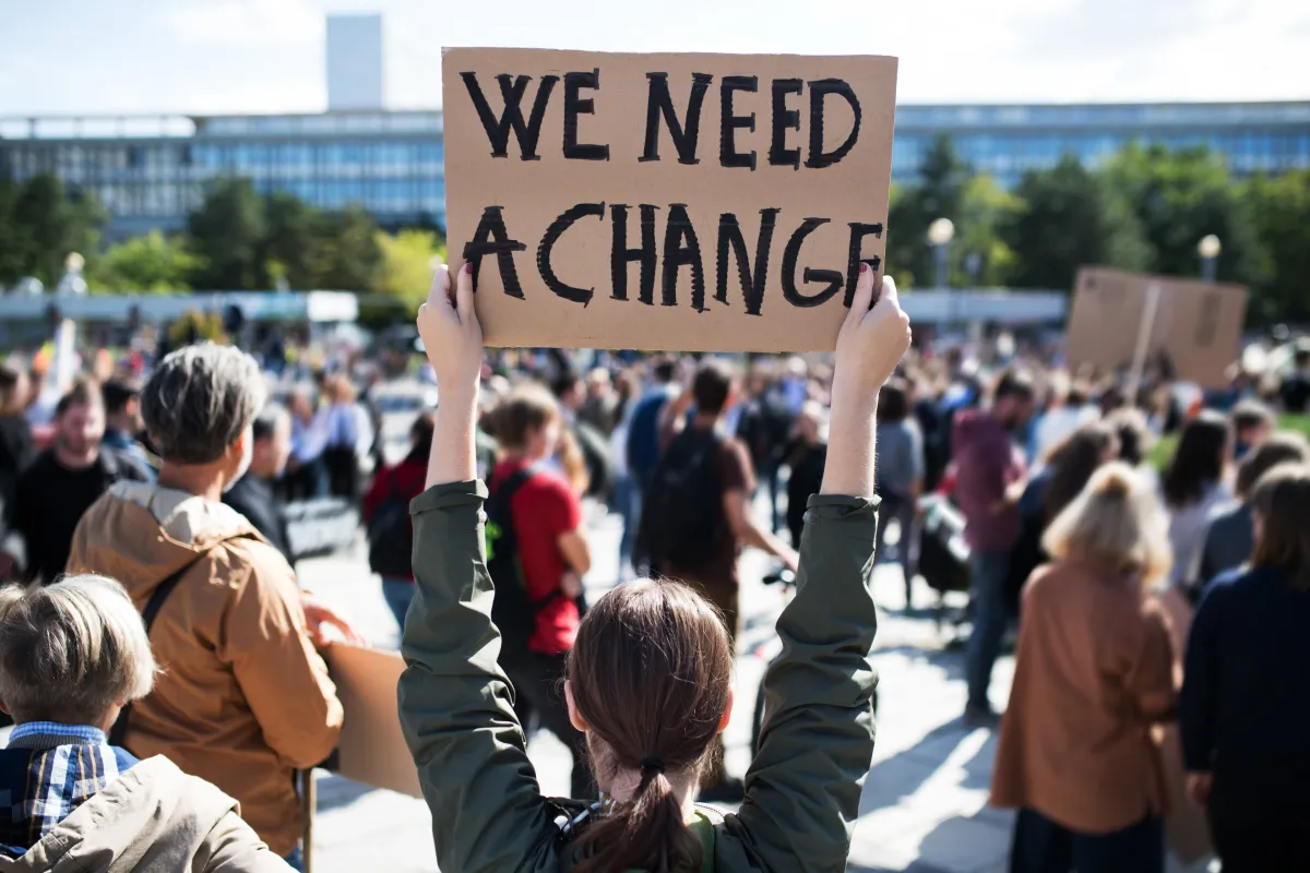 Blick auf eine Protestveranstaltung, viele Menschen von hinten, mittig steht eine Frau und hält ein Plakat mit der Aufschrift "We need a change" hoch